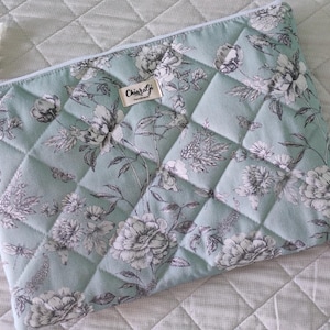 Op de afbeelding: Gewatteerde etui in zachtgroene kleur, versierd met een bloemenpatroon in grijstinten en wit. De etui heeft een ruitvormig stikselontwerp en een klein label met de tekst "Chiaralgi handmade". Een witte koordlus is aan de rits bevestigd.