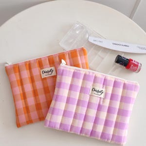 May include: Two quilted fabric pouches in orange and lavender gingham patterns. Each pouch has a zipper closure and a small white label that reads "ChiaralGi Handmade". A red nail polish bottle, nail file, and nail tips are also visible.