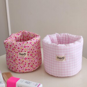 Op de afbeelding: Twee stoffen opbergcontainers. Eén is roze met een bloemenpatroon, de andere is roze en wit geruit. Beide hebben een label met de tekst "Chiaragi Handmade". De containers zijn cilindrisch en staan op een wit oppervlak.