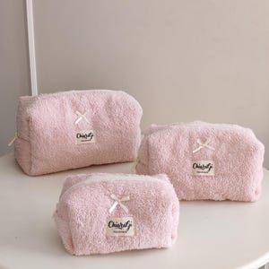 May include: Three light pink, textured, terry cloth cosmetic bags of varying sizes. Each bag features a small white ribbon bow above a rectangular fabric label with the text "Chiaragi Handmade" in black script and a white zipper closure.