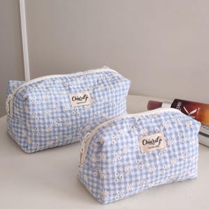 Puede incluir: Dos estuches rectangulares con cremallera que presentan un patrón de cuadros vichy en azul claro y blanco con detalles florales bordados en blanco. Cada estuche tiene una etiqueta de tela con el texto ChiaraGi Handmade en negro.