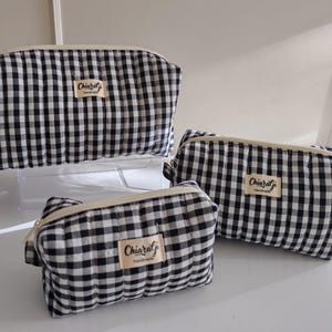 Op de afbeelding: Drie zwart-wit geruite make-uptassen in verschillende maten. Elke tas heeft een beige rits en een klein rechthoekig label met de tekst "ChiaraGi Handmade". De tassen zijn geplaatst op een wit oppervlak.