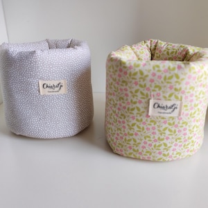Op de afbeelding: Twee stoffen containers. Eén is lichtgrijs met een wit stippenpatroon. De andere is wit met een bloemenpatroon van roze bloemen en groene bladeren. Elk heeft een label met "Chiaralgi Handmade".