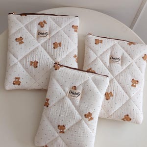 May include: Three quilted pouches in off-white fabric with a brown bear and small dot pattern. Each rectangular pouch features a brown zipper closure and a beige fabric label with the text ChiaraGi Handmade stitched on the front surface.
