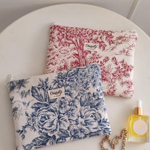 May include: Two rectangular cosmetic bags with zipper closures. One bag features a red and white toile pattern, and the other has a blue floral design. The bags are labeled "ChiaraGi Handmade". A gold necklace and a small bottle of oil are also present.