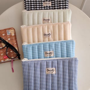 May include: Five quilted pencil cases in a row, each with a different gingham pattern. The colors include black and white, cream, peach, light blue, and navy blue. Each case has a zipper and a small label that reads "Chiaragi Handmade."