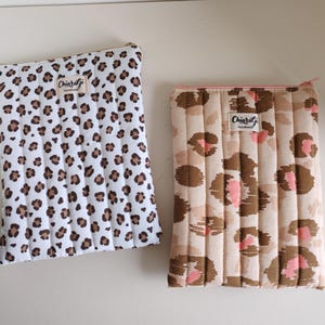 Könnte beinhalten: Zwei gesteppte Stoffhüllen mit Reißverschlüssen. Eine Hülle ist weiß mit braunen Leopardenflecken, die andere beige mit rosa und braunen Leopardenflecken. Jede Hülle hat ein kleines Etikett mit der Aufschrift "Chiaragi Handmade".
