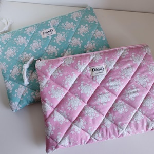 Può includere: Due pochette in tessuto trapuntato, una turchese e una rosa, con un motivo floreale. Ogni pochette ha un design trapuntato diagonale, una chiusura con cerniera e una piccola etichetta con la scritta "Chiaralj". La pochette turchese ha un tirazip a cordino bianco.