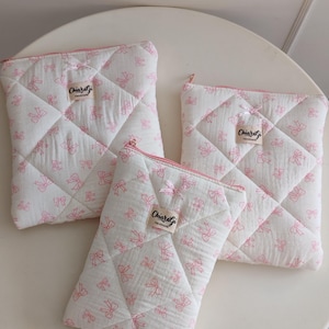 Può includere: Tre pochette trapuntate di diverse dimensioni realizzate in tessuto bianco con un motivo ripetuto di fiocchi rosa. Ogni pochette presenta una cerniera rosa, un piccolo fiocco rosa e un'etichetta in tessuto beige con la scritta ChiaraGi Handmade.