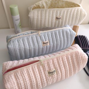 Op de afbeelding: Drie cosmetische tassen in crème, lichtblauw en roze, met een wafeltextuur. Elke tas heeft een ritssluiting en een label met de tekst "Chiaralj Handmade". Een fles Biorevital-spray is zichtbaar. De tassen zijn open en tonen hun binnenkant.