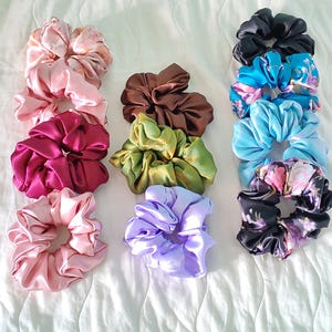 Può includere: Una collezione di elastici per capelli in raso in vari colori, tra cui rosa, bordeaux, marrone, verde, viola, blu e nero. Alcuni elastici presentano motivi floreali. Gli elastici sono disposti su una superficie bianca.