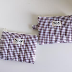 Può includere: Due piccole pochette rettangolari con motivo a quadretti marrone e bianco. Ogni pochette ha una cerniera bianca e un piccolo passante in tessuto. L'etichetta "Chiaragi Handmade" è cucita su ogni pochette.