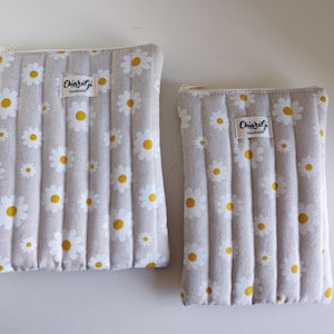 Könnte beinhalten: Zwei beige gesteppte Beutel mit einem Gänseblümchen-Blumenmuster. Die Gänseblümchen haben weiße Blütenblätter und gelbe Zentren. Jeder Beutel hat einen Reißverschluss und ein kleines Etikett mit der Aufschrift "Chiaragi handmade".