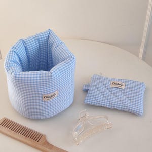 Op de afbeelding: Een opbergmand van lichtblauw en wit gingham-stof en een bijpassend rechthoekig tasje met rits, beide voorzien van een klein wit label met de tekst ChiaraGi. Ook inbegrepen zijn een doorzichtige haarklem en een houten kam.