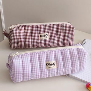 Pode incluir: Dois estojos acolchoados com padrão xadrez vichy, um bordô e branco, o outro lavanda e branco. Cada estojo tem fecho de correr e uma etiqueta com a inscrição "ChiaralGi Handmade". Um caderno e uma caneta são visíveis.