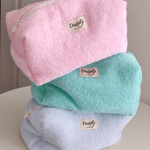 Può includere: Tre trousse per cosmetici in peluche impilate. La trousse superiore è rosa, quella centrale è turchese e quella inferiore è azzurra. Ogni trousse ha una cerniera bianca e un'etichetta con la scritta "ChiaralGi Handmade".