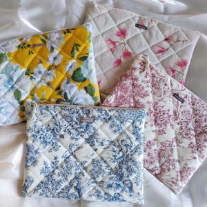 Può includere: Quattro pochette in tessuto trapuntato con diversi motivi floreali. Le pochette sono disposte su uno sfondo in tessuto bianco. I motivi includono fiori gialli e bianchi, fiori rosa e bianchi, fiori blu e bianchi e fiori rossi e bianchi.