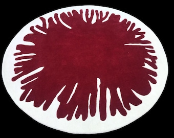 May include: A round, white rug with a large, burgundy-colored design in the center. The design resembles a stylized flower or a burst of energy.