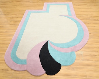 May include: An area rug with an irregular, asymmetrical shape featuring a large cream-colored center section. The rug is bordered by thick bands of light blue and soft pink, with a prominent black teardrop-shaped accent at the bottom edge.
