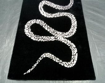 May include: A rectangular black rug featuring a white snake design. The snake is depicted in a winding pattern across the rug's surface. The rug has a soft, textured appearance.