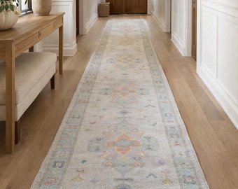 May include: A long, rectangular runner rug featuring a light gray base with a repeating pattern of soft blue, orange, and yellow geometric motifs. The rug has a fringed edge and is positioned centrally along a wooden floor in a hallway.