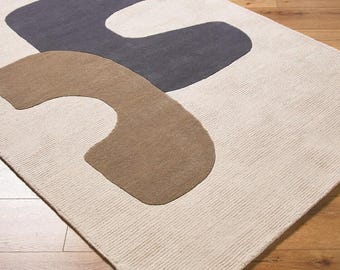 May include: A rectangular area rug with a cream-colored background and two large, abstract shapes in gray and brown. The rug has a textured surface and is placed on a wooden floor.