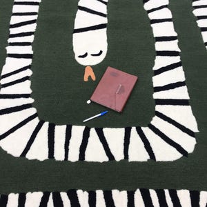 Può includere: Un tappeto verde con un disegno a serpente bianco e nero. Il serpente è avvolto in un motivo curvo. Un taccuino marrone e una penna blu sono sul tappeto.