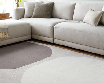 May include: A modern living room featuring a light gray sectional sofa with several throw pillows. A patterned area rug in shades of gray and white is on the floor. A plant and artwork complete the room's decor.