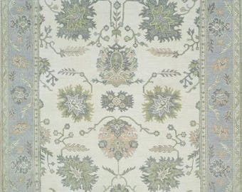 May include: A cream-colored rug with a floral pattern in shades of green, gray, and pink. The rug has a border with a repeating pattern in shades of blue and green.