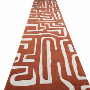 May include: A long, rectangular rug with a terracotta-colored background and a white, abstract pattern. The design features thick, curving lines and shapes, creating a bold, geometric aesthetic. The rug is made of a soft, textured material.