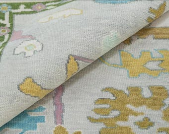 May include: A close-up of a light gray rug with a colorful geometric pattern. The rug features a border with green, pink, and white floral designs. The center of the rug has a repeating pattern of yellow, blue, and pink shapes.