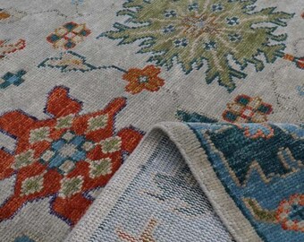 May include: A close-up of a rug with a light gray background and intricate floral patterns in shades of red, orange, blue, and green. The rug has a fringed edge.