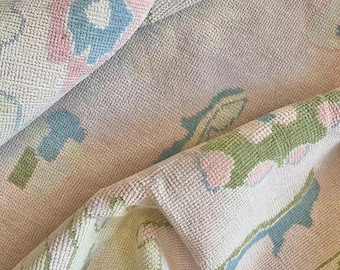 May include: Close-up of a rolled-up, textured rug with a floral pattern. The rug features a light pink background with blue, pink, and green floral designs. The edges have a border with geometric shapes. The rug appears to be made of a soft, woven material.