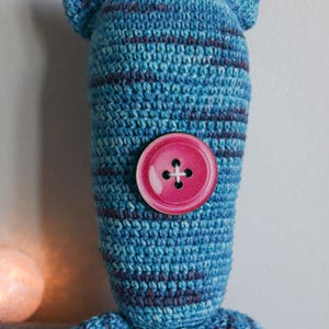 May include: A handmade blue and purple striped crochet doll with a large pink button. The doll has a rounded head and body. A small, glowing, round light is in the background.