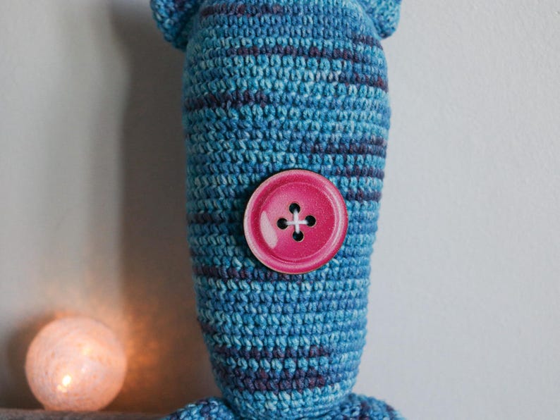 May include: A handmade blue and purple striped crochet doll with a large pink button. The doll has a rounded head and body. A small, glowing, round light is in the background.