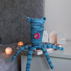 May include: A handmade blue and navy striped crochet octopus toy with a pink button and small ears. The toy is sitting on a book, with decorative lights and small white house-shaped candle holders in the background.