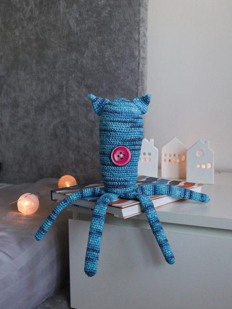 May include: A handmade blue and navy striped crochet octopus toy with a pink button and small ears. The toy is sitting on a book, with decorative lights and small white house-shaped candle holders in the background.