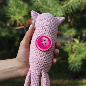 May include: A handmade, light pink crocheted octopus toy with a large, bright pink button detail. The octopus has small ears and tentacles. The toy is held against a backdrop of green foliage.