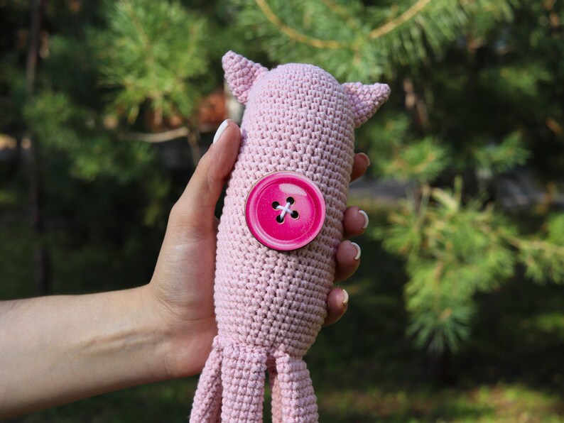 May include: A handmade, light pink crocheted octopus toy with a large, bright pink button detail. The octopus has small ears and tentacles. The toy is held against a backdrop of green foliage.
