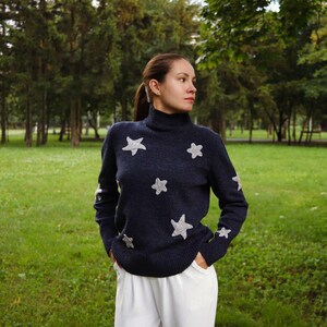 May include: Navy blue turtleneck sweater with silver star appliqu&eacute;s. The long-sleeved sweater has stars on the front and sleeves. The person is wearing white pants and standing in a grassy park.