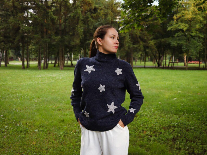 May include: Navy blue turtleneck sweater with silver star appliqu&eacute;s. The long-sleeved sweater has stars on the front and sleeves. The person is wearing white pants and standing in a grassy park.