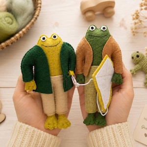 Könnte beinhalten: Zwei Froschpuppen aus Filz, eine gelb mit dunkelgrüner Jacke und beiger Hose, die andere grün mit brauner Jacke und hellgrüner Hose, die einen weißen Drachen halten. Die Puppen haben aufgestickte Augen und einfache Gliedmaßen aus weichem Filz.