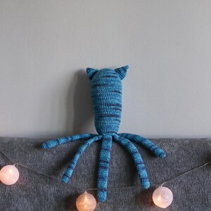 May include: A handmade blue and navy striped crochet octopus plush toy with pointed ears and eight long tentacles. The plush is resting on a gray surface, with a string of glowing white spherical lights in the foreground.