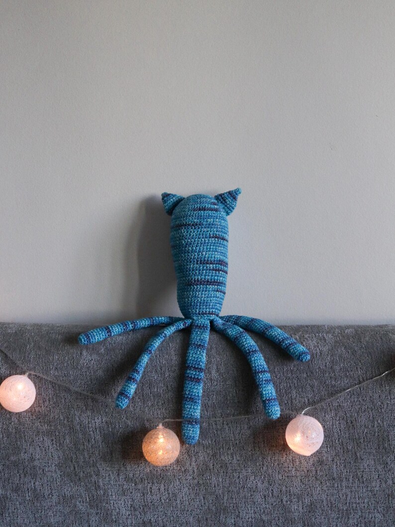 May include: A handmade blue and navy striped crochet octopus plush toy with pointed ears and eight long tentacles. The plush is resting on a gray surface, with a string of glowing white spherical lights in the foreground.