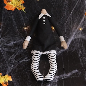 May include: A headless doll dressed in a black dress with a white collar and cuffs. The doll has striped black and white stockings and tan hands. The doll is decorated with white lace trim and three white buttons. The background is a dark surface with a spiderweb design and fall leaves.