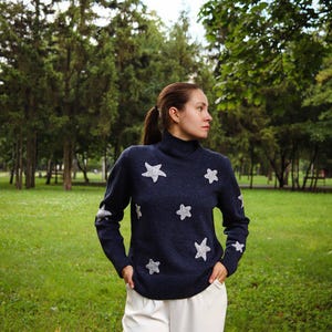 May include: A navy blue turtleneck sweater with white star patterns. The sweater is paired with white pants. The image is taken outdoors with a green grassy background and trees.