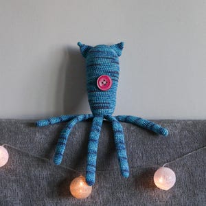May include: A handmade blue and teal crochet creature with a pink button eye and four tentacles. The creature has pointed ears and is resting on a gray surface. A string of glowing white and orange fairy lights is in the foreground.