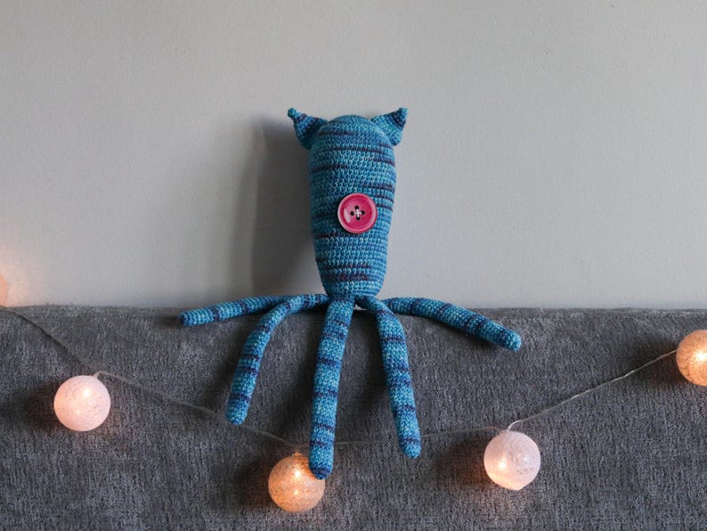 May include: A handmade blue and teal crochet creature with a pink button eye and four tentacles. The creature has pointed ears and is resting on a gray surface. A string of glowing white and orange fairy lights is in the foreground.