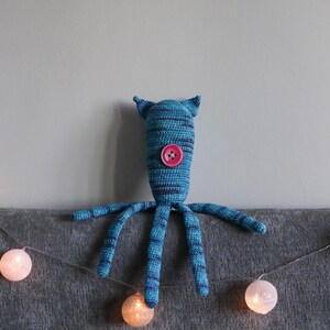 May include: A handmade blue and navy striped crochet monster toy with a pink button and small ears. The toy has six long, striped arms. A string of glowing, round, white and orange lights is in the foreground.