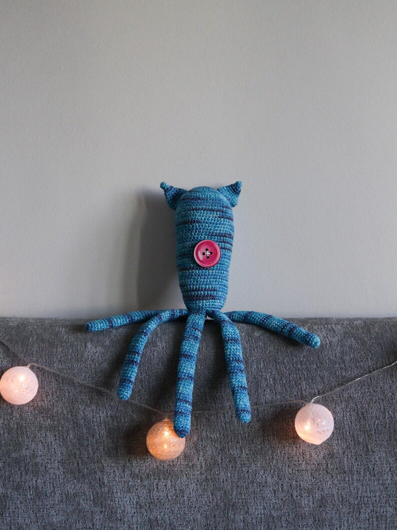 May include: A handmade blue and navy striped crochet monster toy with a pink button and small ears. The toy has six long, striped arms. A string of glowing, round, white and orange lights is in the foreground.